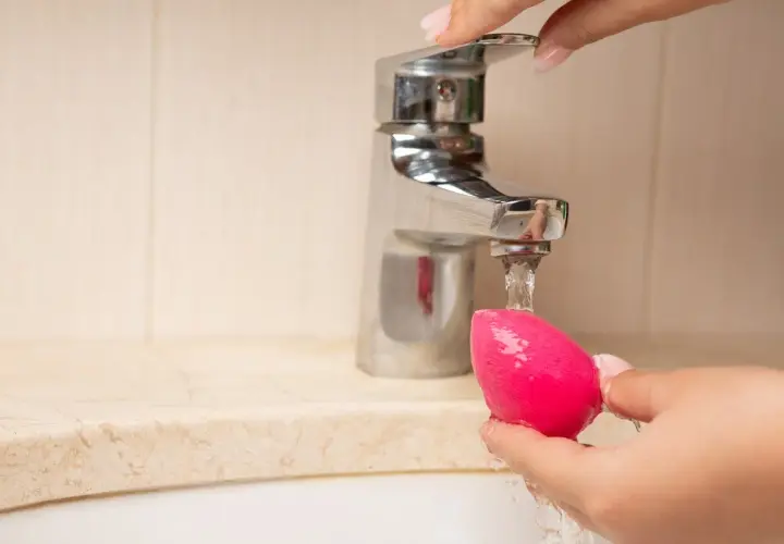 Mano limpiando esponja de maquillaje rosa bajo un grifo. Aprende como limpiar las esponjas de maquillaje para un mejor acabado.