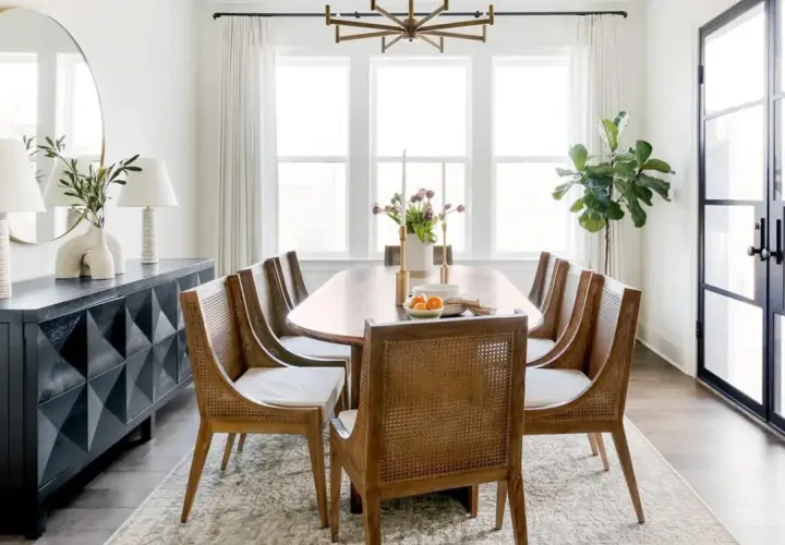 A spacious dining room with a large oval table, cane-backed chairs, and a modern sideboard. The dining room width is emphasized by the large windows and glass doors.
