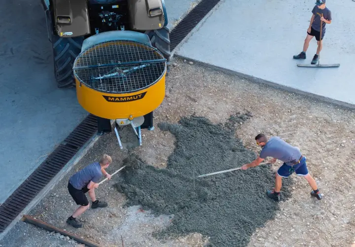 Zwei Arbeiter verteilen frischen Beton, der aus einem gelben Zwangsmischer einer Mammut-Maschine fließt. Ein dritter Arbeiter glättet den Beton.