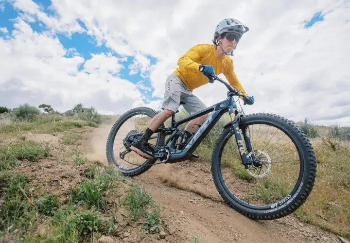 A mountain biker shreds a dusty trail on a Yeti MTE bike, kicking up dirt under a cloudy sky.