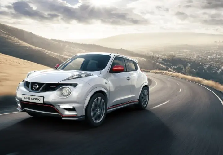 Biały Nissan Juke Nismo z czerwonymi lusterkami pędzi po krętej drodze z widokiem na miasto w oddali.