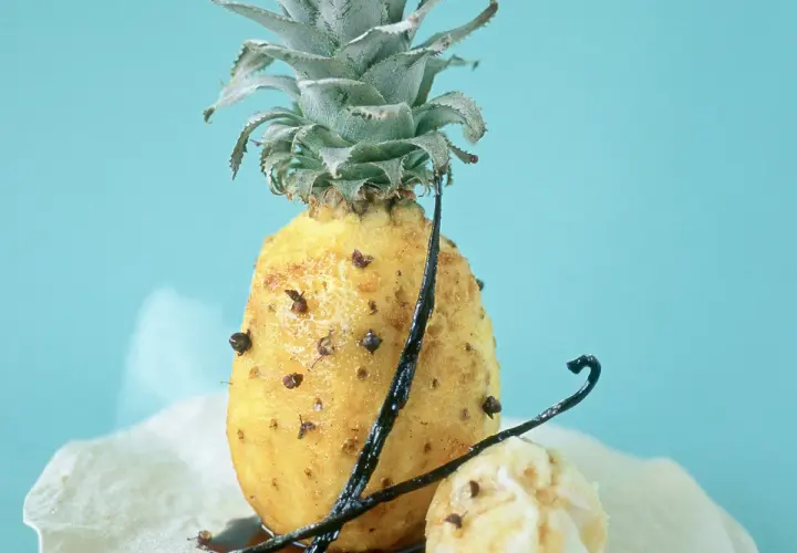 Un ananas rôti, parsemé de clous de girofle et de gousses de vanille, sur un fond bleu pâle. Un délice sucré et épicé.