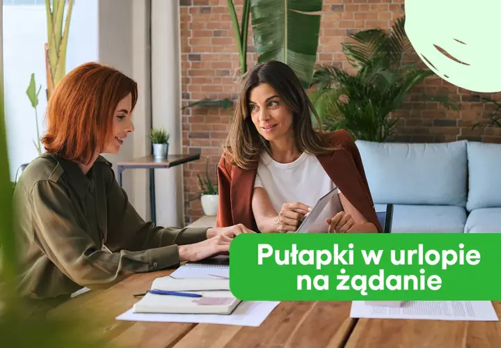 Dwie kobiety rozmawiają przy biurku, jedna pokazuje coś na tablecie.