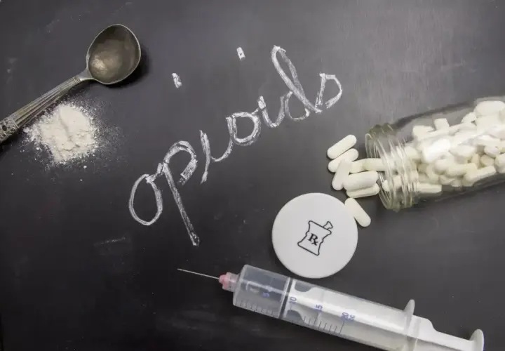Na czarnej tablicy napis "opioids" kredą. Obok łyżka z proszkiem, strzykawka, fiolka z kapsułkami i symbol apteczny.