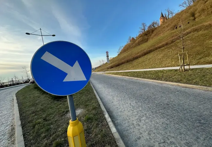 Niebieski znak drogowy ze strzałką w prawo na tle zielonego wzgórza i nieba.
