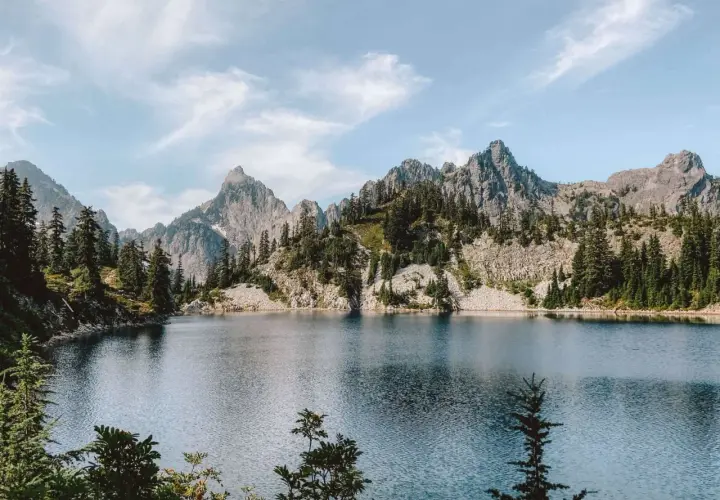 A serene alpine lake reflects jagged, tree-covered mountains. This view makes you wonder how long to hike 16 miles to reach such a peaceful spot.