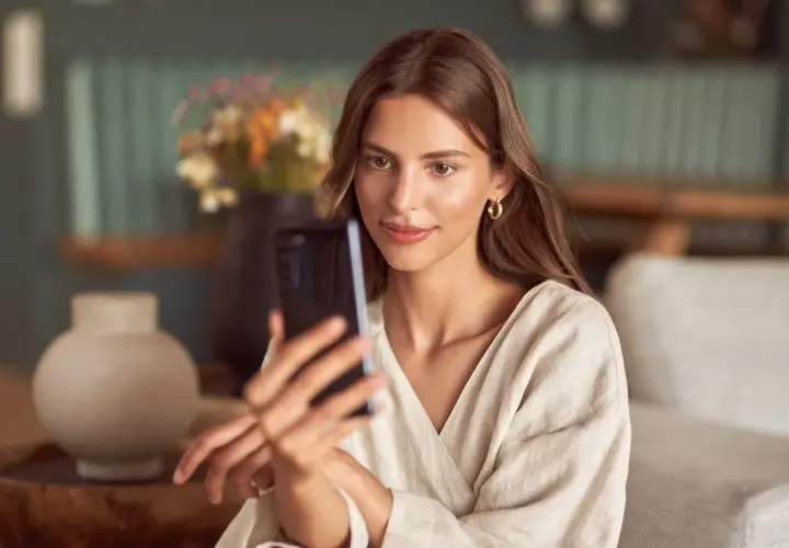 Kobieta robi sobie selfie, patrząc na ekran telefonu. Delikatne światło podkreśla jej urodę, a subtelny uśmiech sugeruje, że wie, jak zrobić dobre selfie.