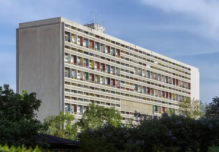 Wielopiętrowy budynek z kolorowymi balkonami, symbol epoki modernizmu, otoczony zielenią.