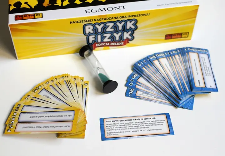 Pudełko gry "Ryzyk Fizyk" i rozłożone karty pytań i odpowiedzi. Czasomierz piaskowy czeka na rozpoczęcie rundy.