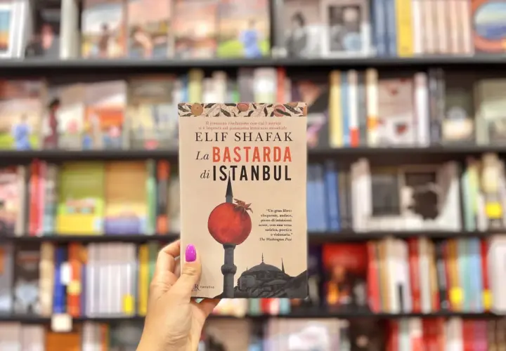 Mano tiene "La Bastarda di Istanbul", un libro che esplora le storie di Istanbul, tra scaffali pieni di libri.