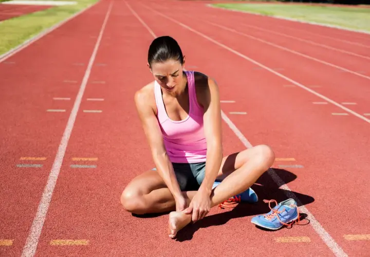 Biegaczka siedzi na bieżni, masując bolącą piętę po treningu.