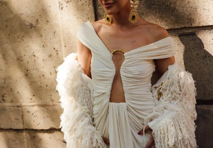Elegancka sesja fashion: modelka w kremowej sukni z wycięciem i futrzanym bolerkiem, ozdobiona złotą biżuterią.