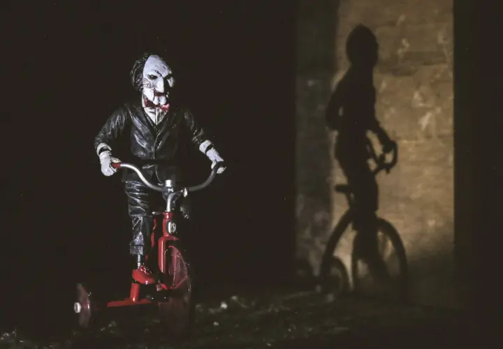 Lalka Billy z Piły na czerwonym trójkołowcu, przypominająca scenę z najlepszych horrorów. Cień na ścianie dodaje grozy.