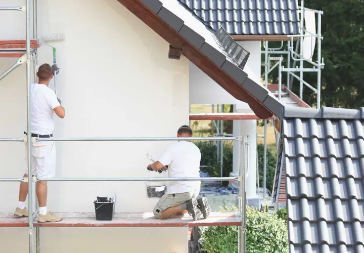 Dwóch malarzy na rusztowaniu odnawia elewację domu. Używają wałków i wiadra z farbą malarską rozpuszczalną w wodzie.