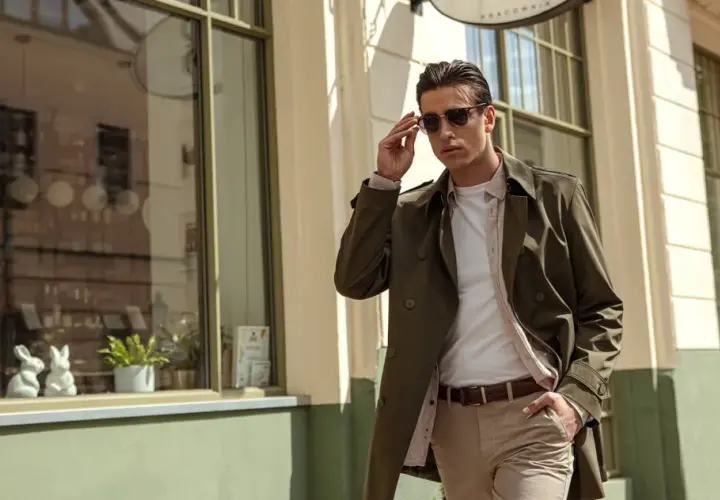 Mężczyzna w eleganckim, khaki płaszczu i beżowych spodniach poprawia okulary, idąc ulicą.
