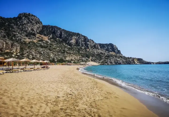 Piękna, piaszczysta plaża Tsambika z turkusową wodą i skalistym wzgórzem w tle. Leżaki i parasole czekają na plażowiczów.