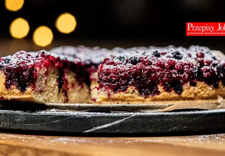 Pyszne ciasto z porzeczkami, posypane cukrem pudrem, na ciemnym talerzu. W tle rozmyte, ciepłe światła.