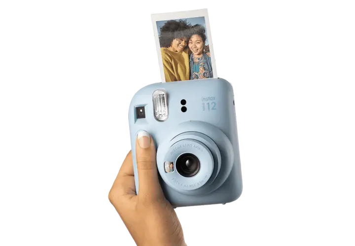 Niebieski aparat Instax Mini 12, trzymany w dłoni, z właśnie wysuniętym, jasnym zdjęciem dwóch uśmiechniętych przyjaciółek.