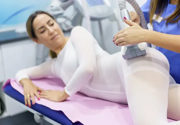 Kobieta w białym kombinezonie leży na kozetce, a kosmetyczka wykonuje zabieg endermologii. To nowoczesny icoon piękna.