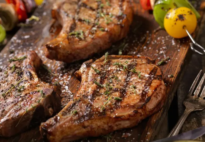 Trois côtelettes de porc grillées, garnies d'herbes et de gros sel, sur une planche en bois. Une surprise de porc barbecue prête à être dégustée.