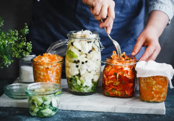 Här är burkar med fermenterad mat: blomkål, morot och kål. En hand använder en gaffel för att plocka från en burk.