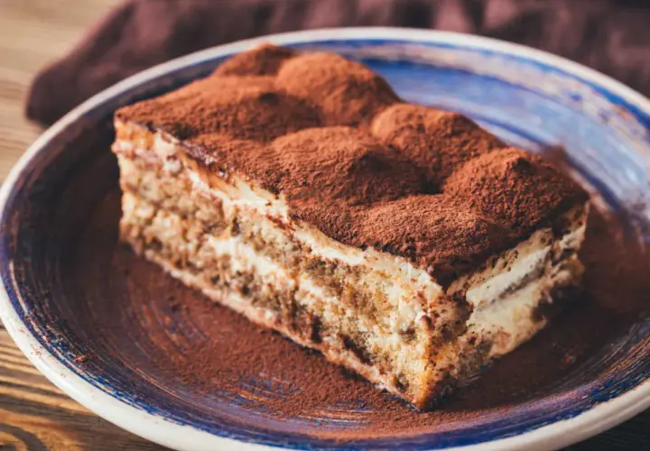 Une part généreuse de tiramisu cake, saupoudrée de cacao, repose sur une assiette bleue. Un délice italien.