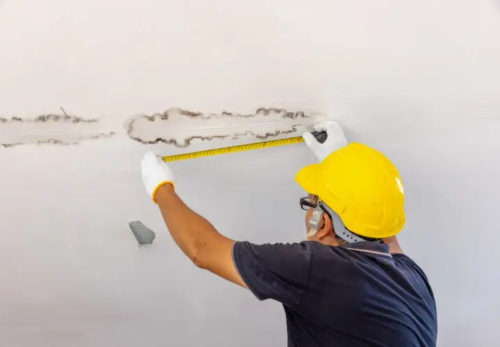 Un homme en casque jaune mesure des taches d'humidité au plafond, cherchant une solution humidité maison.