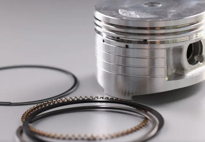 Partes de un pistón: un pistón metálico con ranuras y anillos de compresión y de aceite dispuestos a su lado.