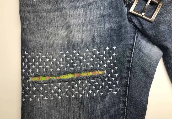 Close-up of denim jeans with a colorful fabric patch and white cross-stitch embroidery, showcasing a stylish sashiko jeans repair.