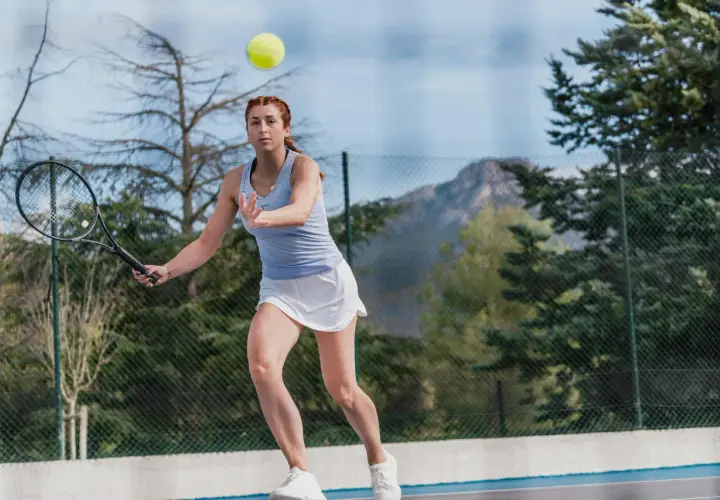 Kobieta w białej spódniczce i niebieskiej koszulce ćwiczy tenis przy ścianie, odbijając piłkę rakietą.