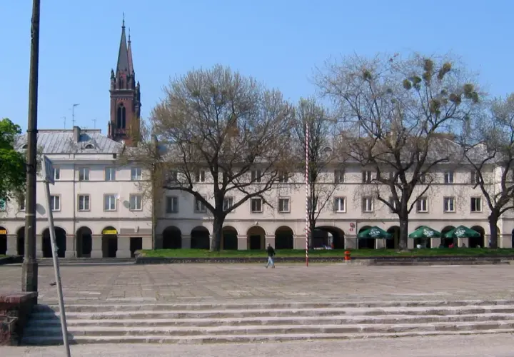 Łódź rynek z kamienicami, drzewami i wieżą kościelną w tle.