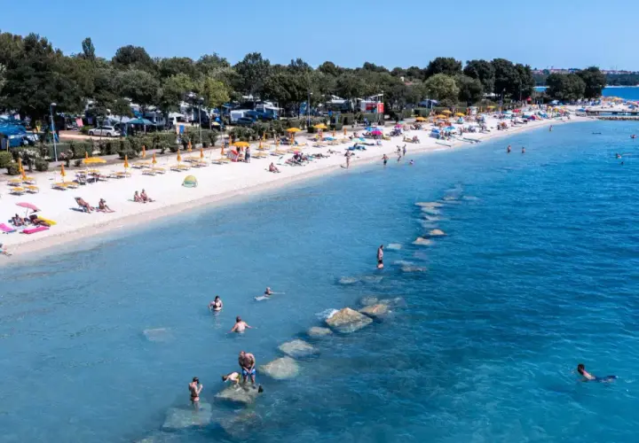 Słoneczna plaża z turystami, parasolami i kamperami w tle. Woda kusi do kąpieli, a kamienie tworzą naturalne falochrony. Idealne miejsce na bi village camping.
