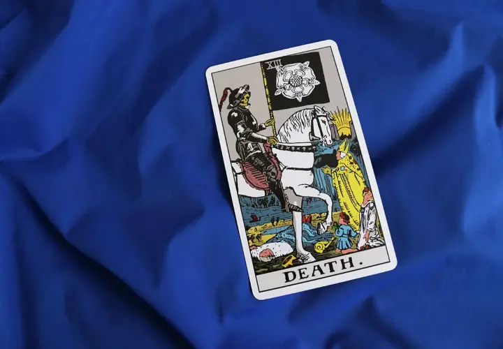 Tarot karta Śmierci na niebieskim tle. Postać w zbroi na białym koniu, niosąca flagę z różą, symbolizuje nieuchronne zmiany i transformację.