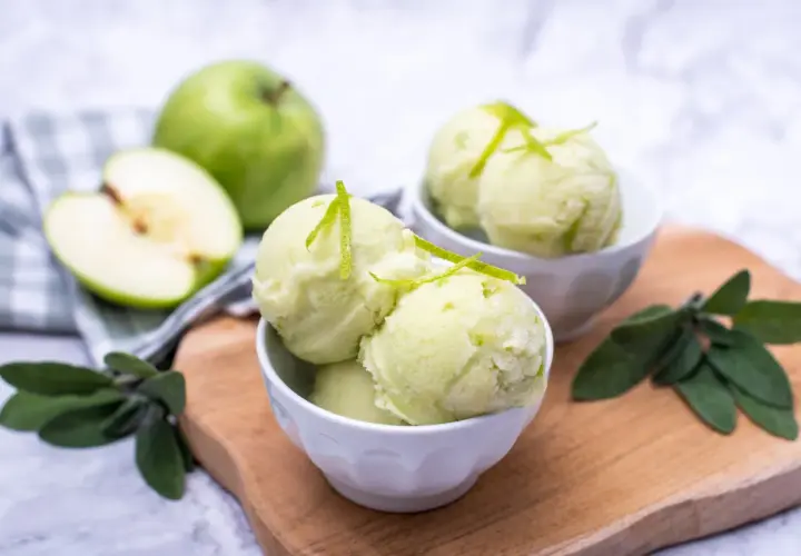 Orzeźwiający sorbet jabłkowy w miseczkach, udekorowany skórką z limonki. Obok całe i połówka zielonego jabłka.