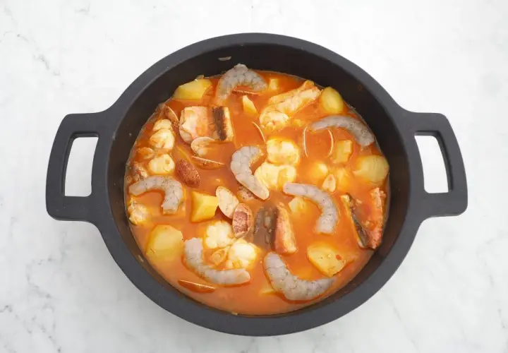 Un delicioso guiso marinero burbujea en una cazuela negra, con patatas, gambas, pescado y almejas en una salsa rojiza.