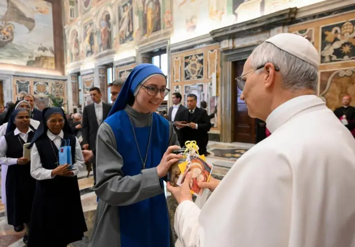 Eine Ordensfrau überreicht dem Papst ein Geschenk. Im Hintergrund stehen weitere Ordensschwestern und Männer in Anzügen.