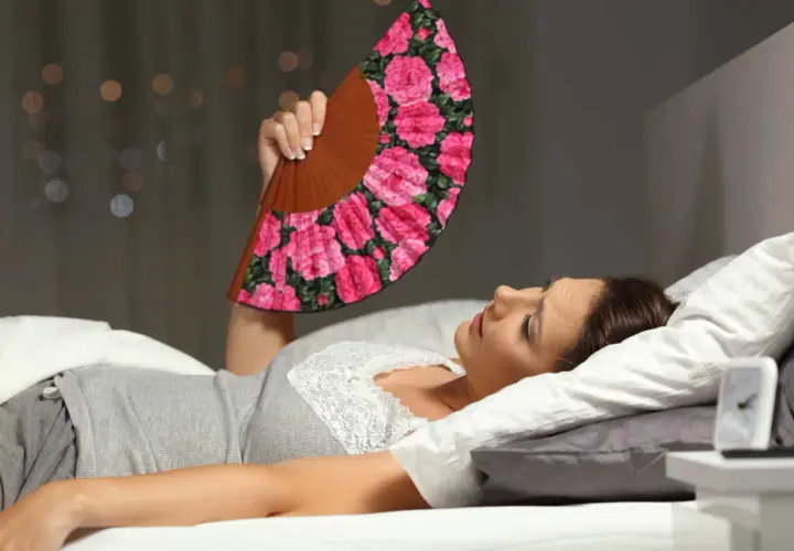 Donna a letto, insonne per il caldo, agita un ventaglio fiorato per combattere la sudorazione notturna.