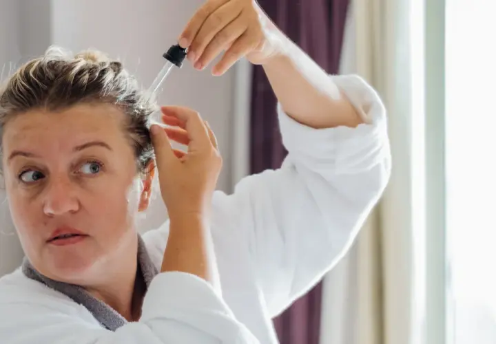 Kobieta w szlafroku aplikuje serum na skórę głowy. Wierzy, że goździki i rozmaryn na włosy przyniosą jej ulgę i wzmocnią kosmyki.