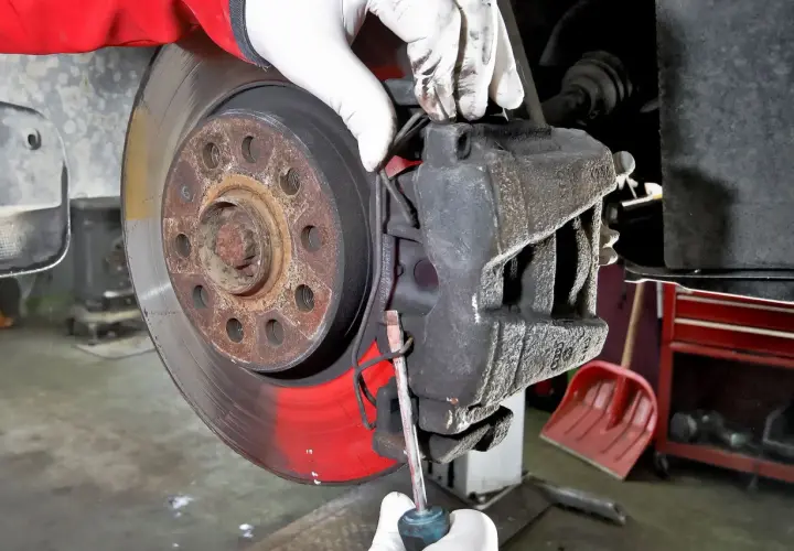 Mechanik w białych rękawiczkach dokonuje wymiany klocków hamulcowych, używając śrubokręta do podważenia starego klocka.