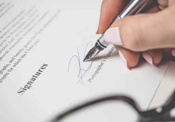 Dłoń z paznokciami malowanymi na różowo podpisuje dokument piórem. Widoczny napis "Signatures" i "Photographer's Signature". To ważny moment, jak wypowiedzenie po angielsku.