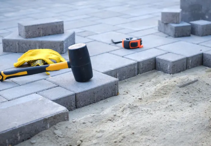 Młotek, rękawice i miarka czekają na ułożenie kostki. W tle widać przygotowany pod nią piasek, a nie beton.