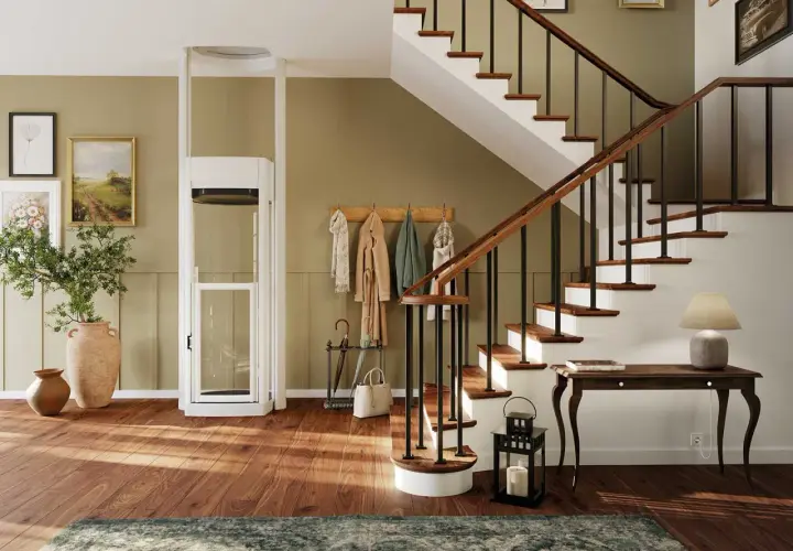 Ein moderner Treppenlift mit Glaskabine steht neben einer Holztreppe. Bilder und Garderobe schmücken die Wand.