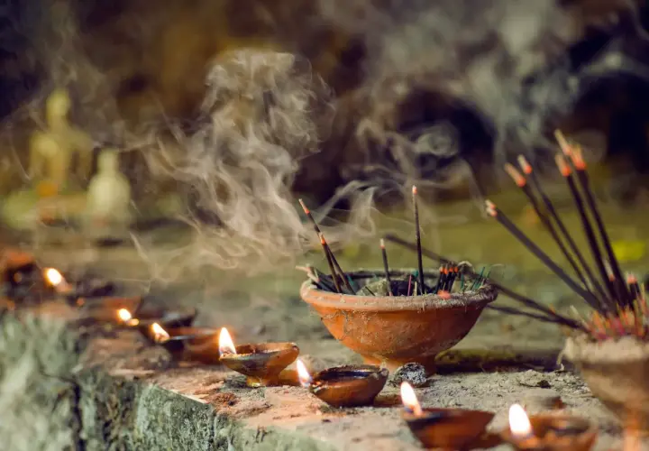 Humo de incienso llenando el aire, un recordatorio de que el incienso es malo para los pulmones. Velas encendidas y estatuillas borrosas en el fondo.