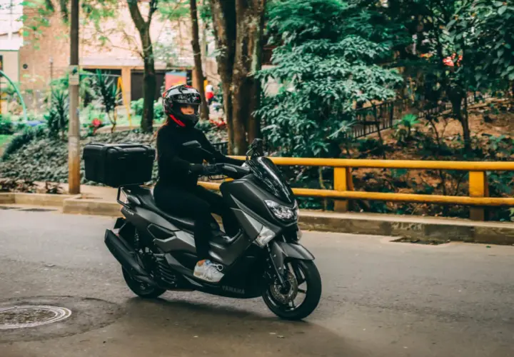 Mujer con casco y ropa negra conduce una moto Yamaha sin marchas, con caja trasera, por una calle arbolada.