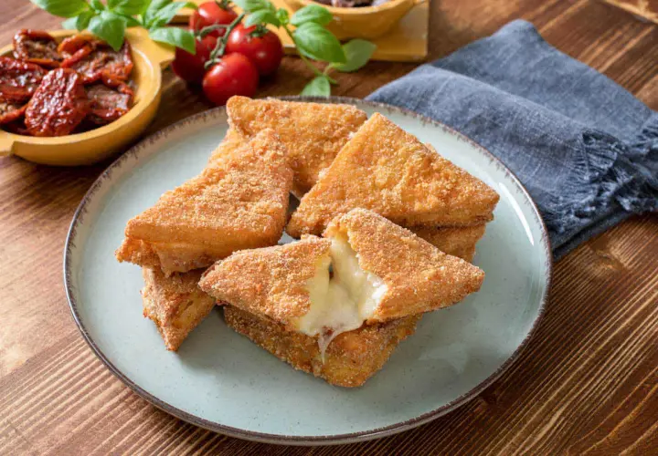 Chrupiące mozzarella in carrozza, z rozpływającym się serem w środku, na talerzu. Obok pomidorki i suszone pomidory.