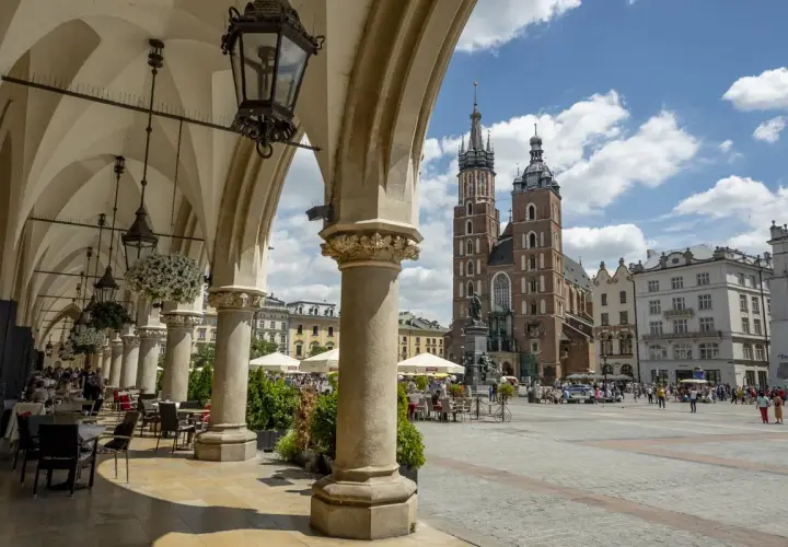 Urokliwy **Kraków Stary Miasto** z Sukiennicami i Kościołem Mariackim w tle. Widać kawiarniane stoliki pod arkadami.