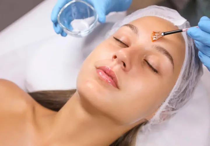 Manos enguantadas aplican un peeling facial con brocha a una mujer con los ojos cerrados.