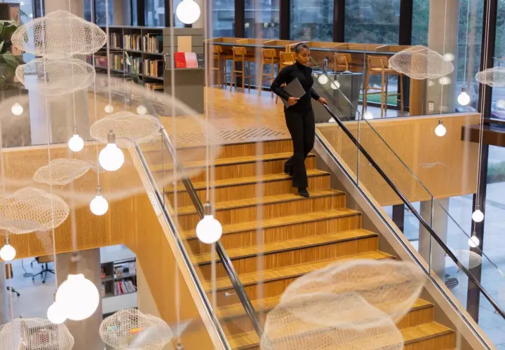Kobieta z laptopem schodzi po drewnianych schodach w nowoczesnej bibliotece. Zastanawia się, ile trwają studia, gdy mija wiszące dekoracje w kształcie chmur.