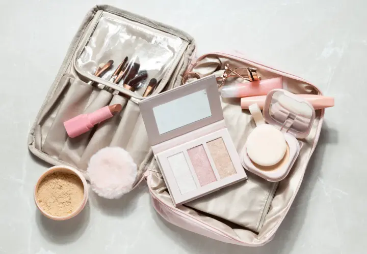 Une trousse de maquillage ouverte, prête pour un voyage en bagage cabine, révèle rouges à lèvres, pinceaux, palette et poudrier.