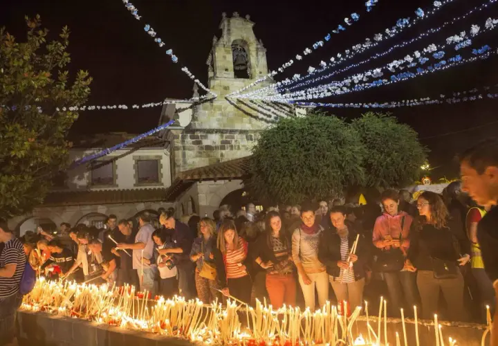 Multitud enciendiendo velas frente a una iglesia iluminada por guirnaldas, parte de las fiestas de Revilla de Camargo.